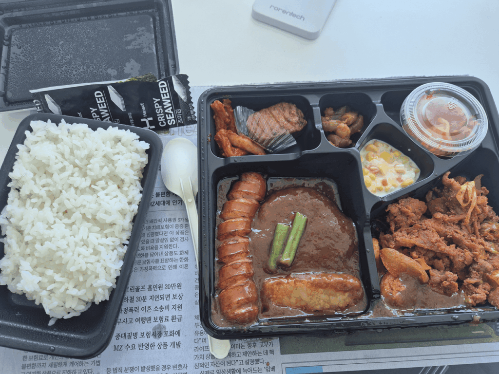 신문지 위에 놓인 1인용 도시락 사진. 한쪽에는 흰 쌀밥이 가득 담겨 있고, 메인 반찬으로는 소시지, 제육볶음, 옥수수 샐러드, 김치 등이 정갈하게 담겨 있다. 도시락 옆에는 포장된 조미김과 일회용 숟가락이 놓여 있어, 바쁜 업무 중 급하게 끼니를 해결하는 사업가의 일상을 보여준다.